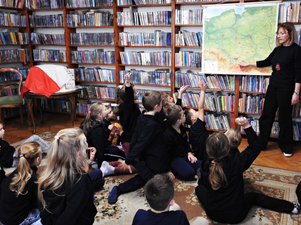 Bibliotekarka wskazuje palcem punkt na mapie Polski. Mapa zawieszona jest na regałach z książkami. Przed bibliotekarką siedzi grupa kilkunastu dzieci. 4 dzieci unosi rękę.  Za dziećmi stolik, na którym znajduje się flaga Polski.