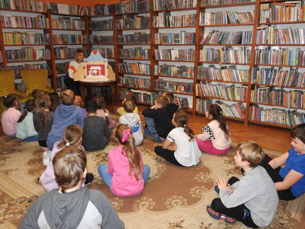 Bibliotekarka siedzi na krześle obok stolika. Na stoliku znajduje się otwarta skrzynia teatrzyku kamishibai. W teatrzyku karta z białymi postaciami na czerwonym tle. Przed skrzynią, na dywanie, siedzi grupa kilkunastu dzieci.  