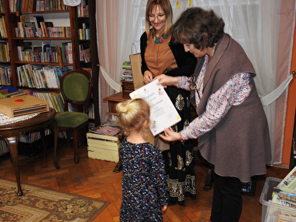 Bibliotekarka wręcza stojącej przed nią dziewczynce dyplom. Obok stoi druga bibliotekarka.