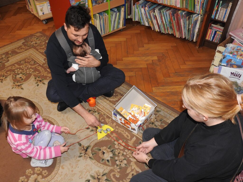 Dwie osoby dorosłe oraz 1 dziecko siedzą na dywanie. Jeden z dorosłych trzyma w ramionach niemowlaka w nosidełku. Przed nimi żółty materiał, na którym leżą szare myszki. Do myszek przyczepione są sznurki. Obok pomarańczowy kubek oraz pudełko do gry.