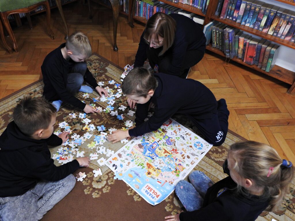 4 dzieci oraz bibliotekarka siedzą wokół plakatu przedstawiającego mapę Polski oraz rozsypanych puzzli. 