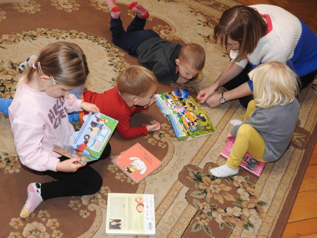 Bibliotekarka oraz 4 dzieci leżą, siedzą na dywanie. Czytają, oglądają książeczki.