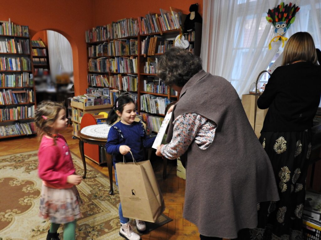 Dziewczynka odbiera dyplom od bibliotekarki. W drugiej ręce trzyma papierową torbę z logo biblioteki.