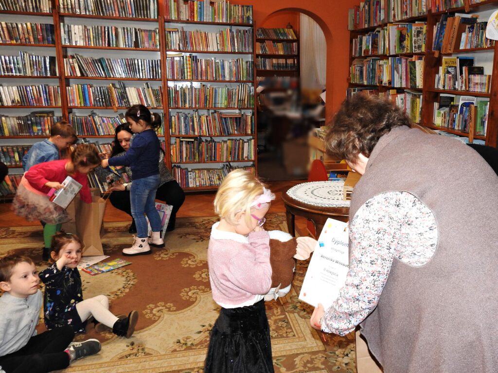 Bibliotekarka wręcza dyplom dziewczynce. Za nimi 5 dzieci oraz 1 osoba dorosła. 2 dzieci siedzi na dywanie, reszta osób zagląda to papierowej torby. W tle regały z książkami.