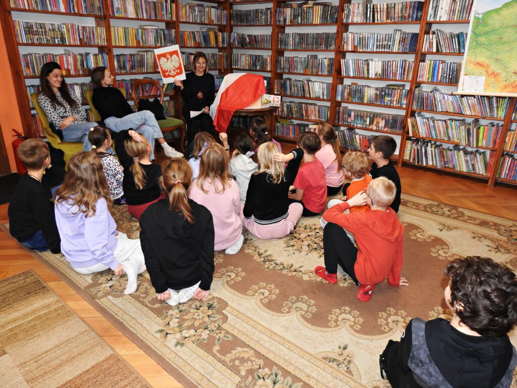 3 osoby dorosłe siedzą na krzesłach. Wśród nich bibliotekarka, która trzyma w uniesionej dłoni kartkę z wydrukiem herbu Polski. Obok niej stolik z flagą Polski. Przed bibliotekarką, na dywanie, siedzi 16 dzieci.