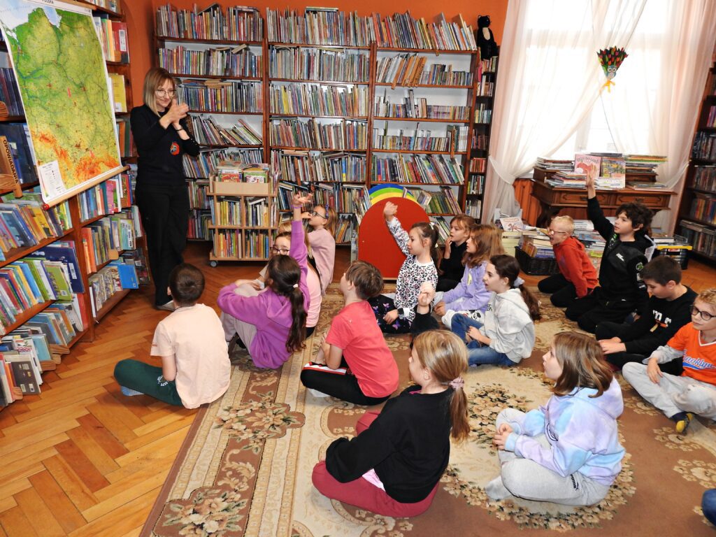 Bibliotekarka stoi przy mapie Polski zawieszonej na regale z książkami. Przed nią, na dywanie siedzi grupa dzieci. 4 dzieci unosi rękę do góry. Bibliotekarka wskazuje dłonią na jedno z dzieci.