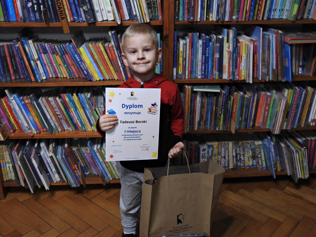 Chłopiec trzyma przed sobą dyplom, w drugiej ręce trzyma papierową torbę z logo biblioteki. Za nim regały z książkami.