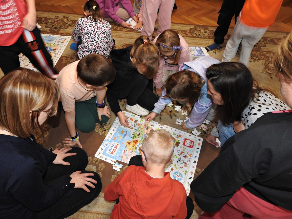 6 dzieci oraz 2 osoby dorosłe są zgromadzone nad puzzlami przedstawiającymi mapę Polski. Puzzle są ułożone, brakuje tylko kilku elementów. 