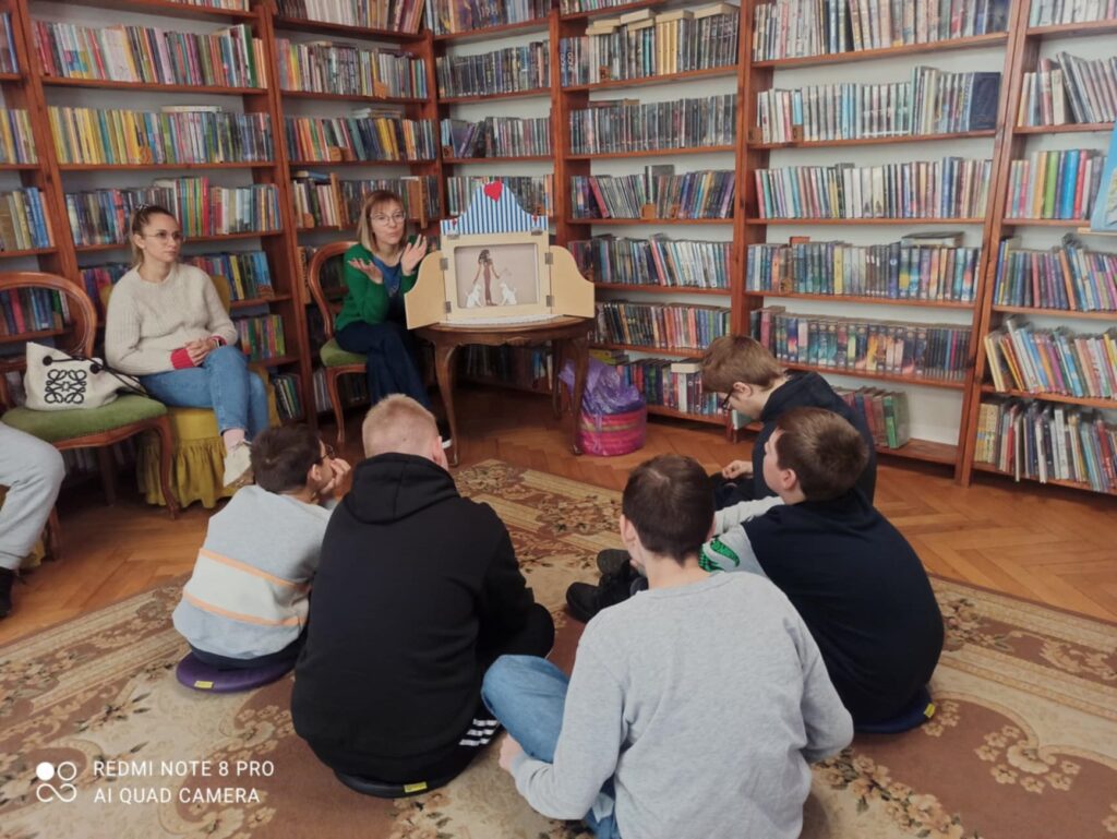 Na dywanie siedzi 5 dzieci. Dzieci patrzą w stronę bibliotekarki, która trzyma w rękach mapę. Bibliotekarka coś pokazuje na mapie.
