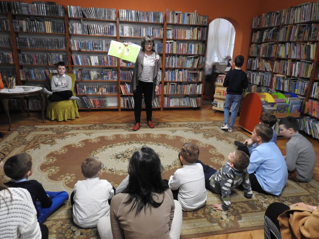 Na dywanie siedzi grupa dzieci. Przed dziećmi stoi bibliotekarka. Bibliotekarka pokazuje ilustrację do książki. 