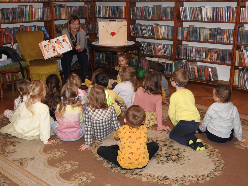 Na dywanie siedzą dzieci i patrzą w stronę siedzącej przed nimi bibliotekarki. Bibliotekarka pokazuje ilustrację z książki. Na ilustracji dzieci z bosymi stopami.