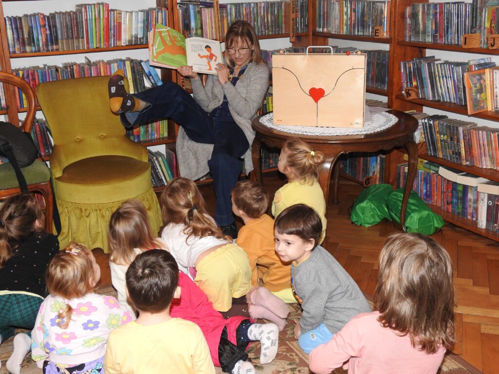 Na dywanie siedzą dzieci i patrzą w stronę siedzącej przed nimi bibliotekarki. Bibliotekarka podnosi nogę  do góry oraz pokazuje ilustrację z książki. Na ilustracji dziecko z bosą stopą oraz małpa.