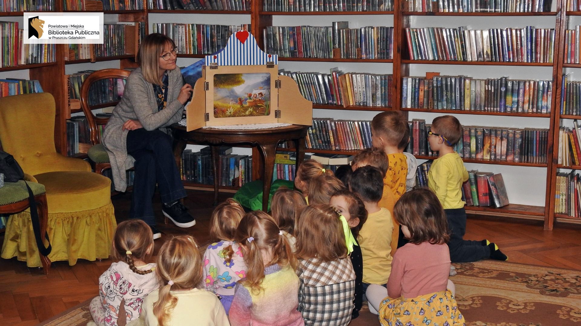 Na dywanie siedzą dzieci. Dzieci patrzą przed siebie, na siedzącą na krześle bibliotekarkę. Obok bibliotekarki stoi stolik, a na nim otwarty teatrzyk kamishibai. Wewnątrz teatrzyku jest ilustracja.