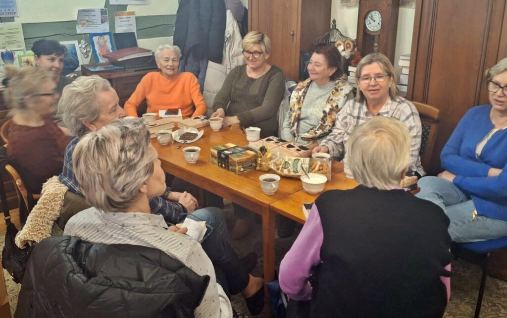 Przy stole siedzą członkinie Dyskusyjnego Klubu Książki. Na stole leżą książki oraz stoją filiżanki z napojem i ciastka.