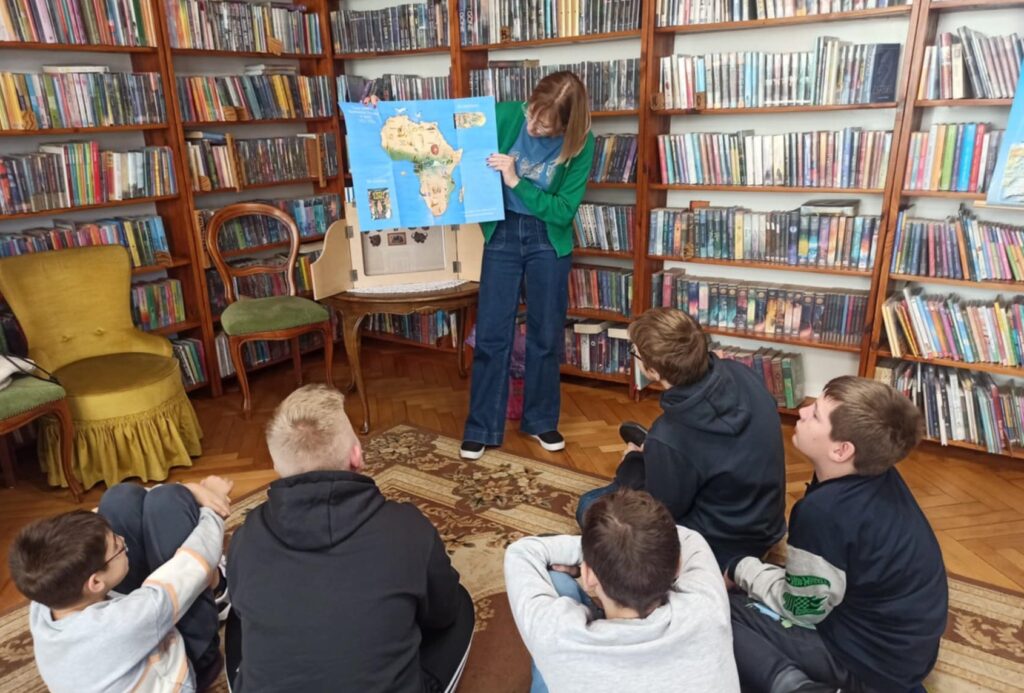 Na dywanie siedzi 5 dzieci. Dzieci patrzą w stronę bibliotekarki, która trzyma w rękach mapę. Bibliotekarka coś pokazuje na mapie.
