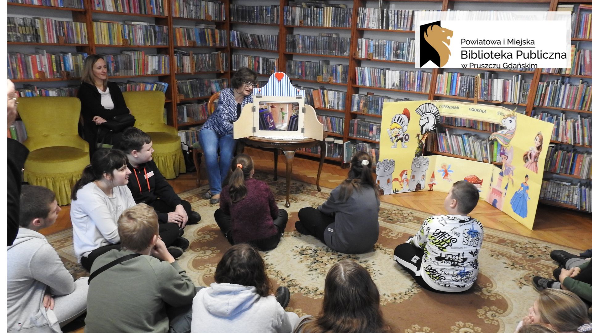 Grupa dzieci siedzi na dywanie i patrzy na siedząca przed nimi bibliotekarkę. Obok bibliotekarki, na stoliku, stoi drewniany teatrzyk kamishibai. Teatrzyk jest otwarty i widać ilustrację. Bibliotekarka czyta.