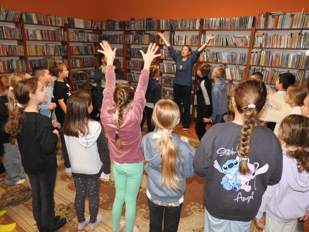 Duża grupa dzieci stoi w sali bibliotecznej. 1 dziecko oraz bibliotekarka trzyma ręce wyciągnięte do góry.