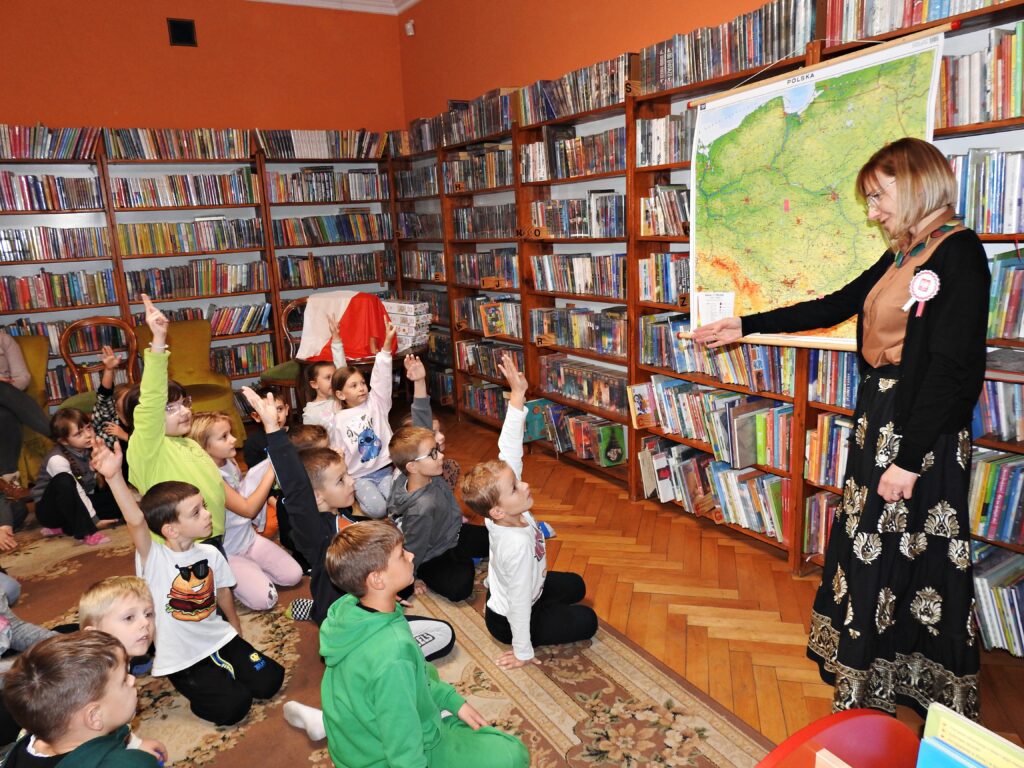 Bibliotekarka stoi przy mapie Polski zawieszonej na regale z książkami. Przed nią, na dywanie siedzi grupa dzieci. 4 dzieci unosi rękę do góry. Bibliotekarka wskazuje dłonią na jedno z dzieci.