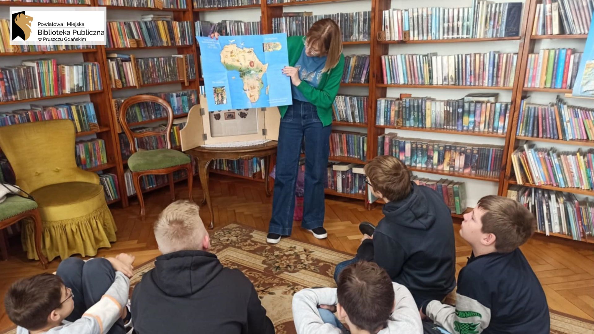 Na dywanie siedzi 5 dzieci. Dzieci patrzą w stronę bibliotekarki, która trzyma w rękach mapę. Bibliotekarka coś pokazuje na mapie.