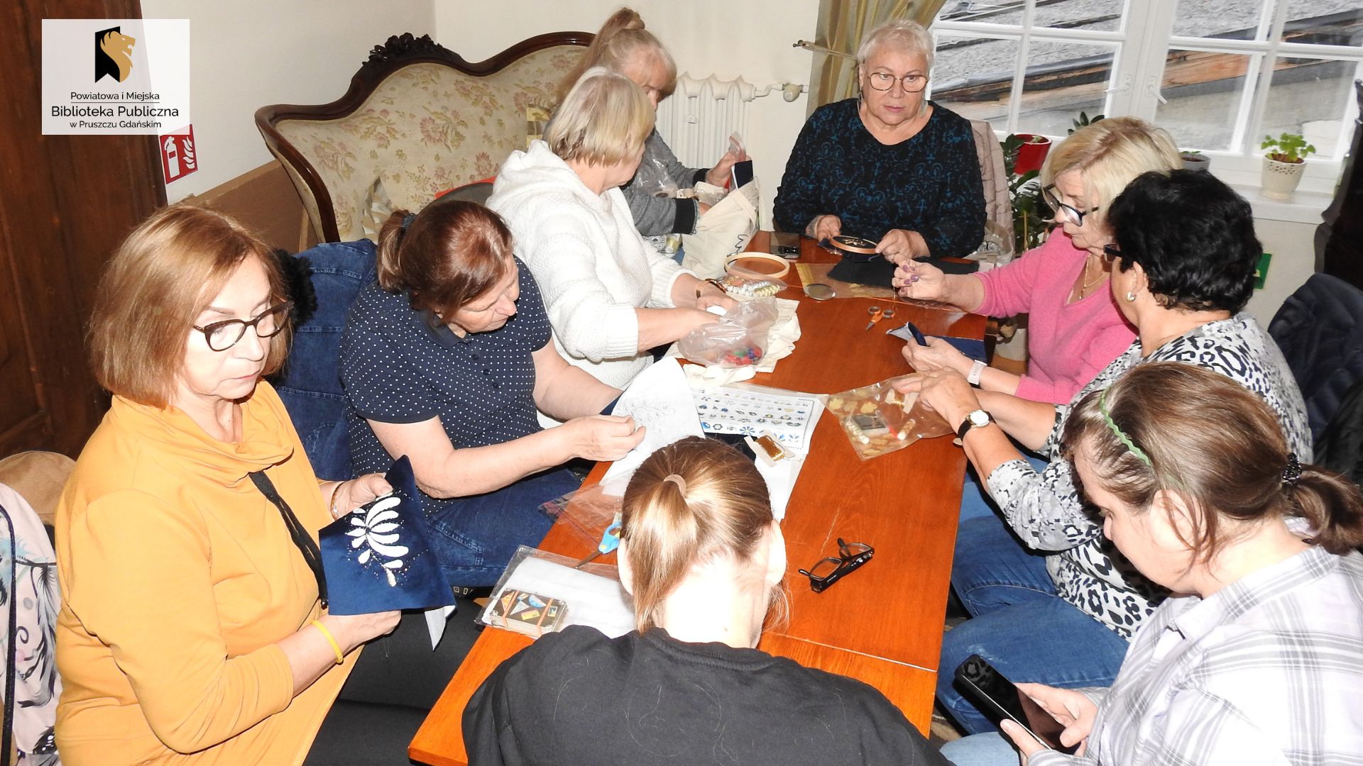 Przy stole siedzi 9 kobiet. Przed kilkoma leżą materiały z częściowo wyhaftowanym wzorem wyhaftowanym wzorem. Inne kobiety haftują.