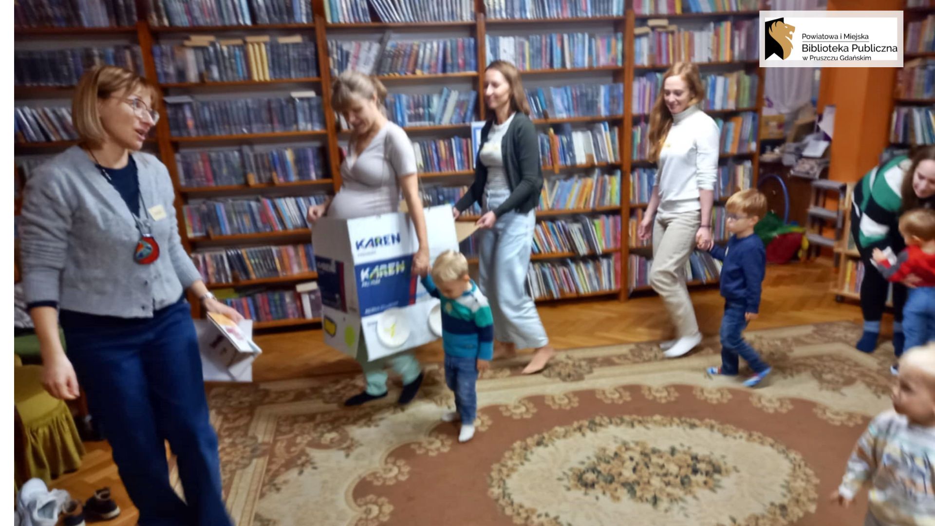 Dzieci oraz dorośli idą jeden za drugim. Na przodzie idzie bibliotekarka z książką w ręku. Za bibliotekarką idzie mama z i trzyma dziecko za rękę. Mama ma założony karton. U dołu kartonu przyklejone są papierowe talerze, które symbolizują koła.