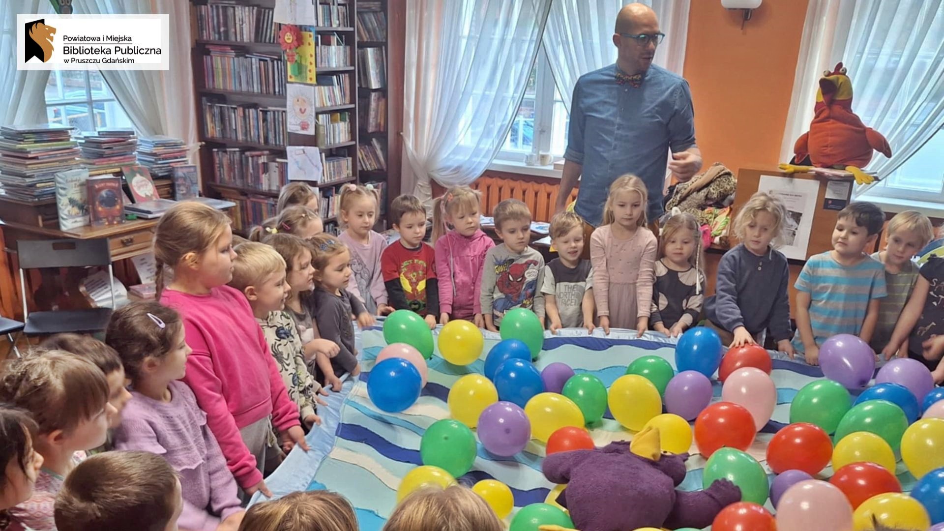 Duża grupa dzieci stoi w okręgu i trzyma za rogi niebieską chustę. Na chuście leżą nadmuchane, kolorowe balony. Obok dzieci stoi gość spotkania — Pan Poeta.