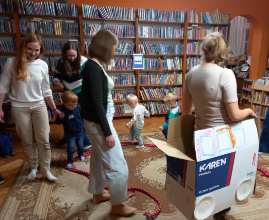 Dzieci oraz dorośli idą jeden za drugim. Na przodzie idzie bibliotekarka z książką w ręku. Za bibliotekarką idzie mama z i trzyma dziecko za rękę. Mama ma założony karton. U dołu kartonu przyklejone są papierowe talerze, które symbolizują koła. 
