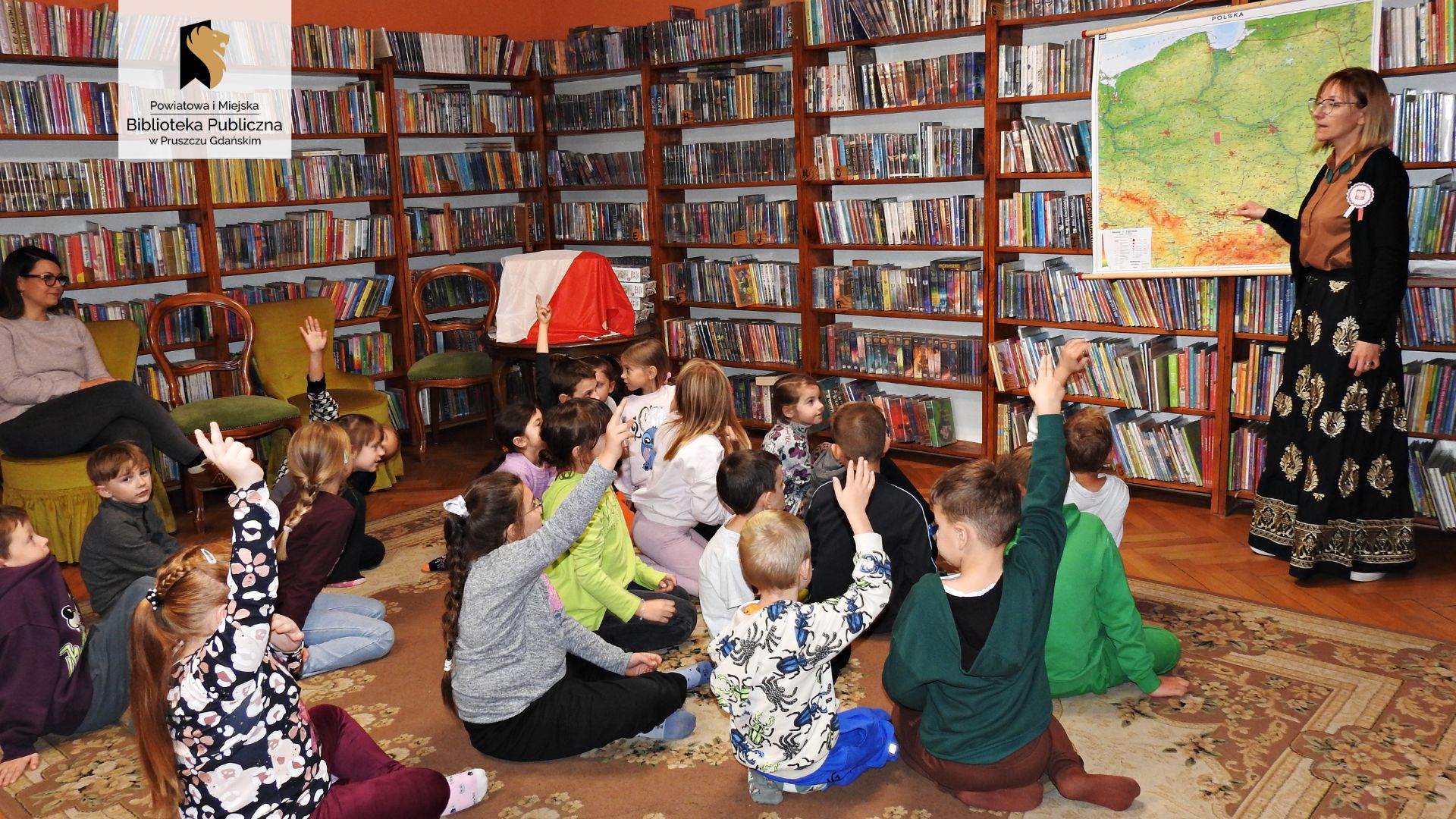 Bibliotekarka wskazuje palcem punkt na mapie Polski. Mapa zawieszona jest na regałach z książkami. Przed bibliotekarką siedzi grupa kilkunastu dzieci. 4 dzieci unosi rękę. Za dziećmi stolik, na którym znajduje się flaga Polski.