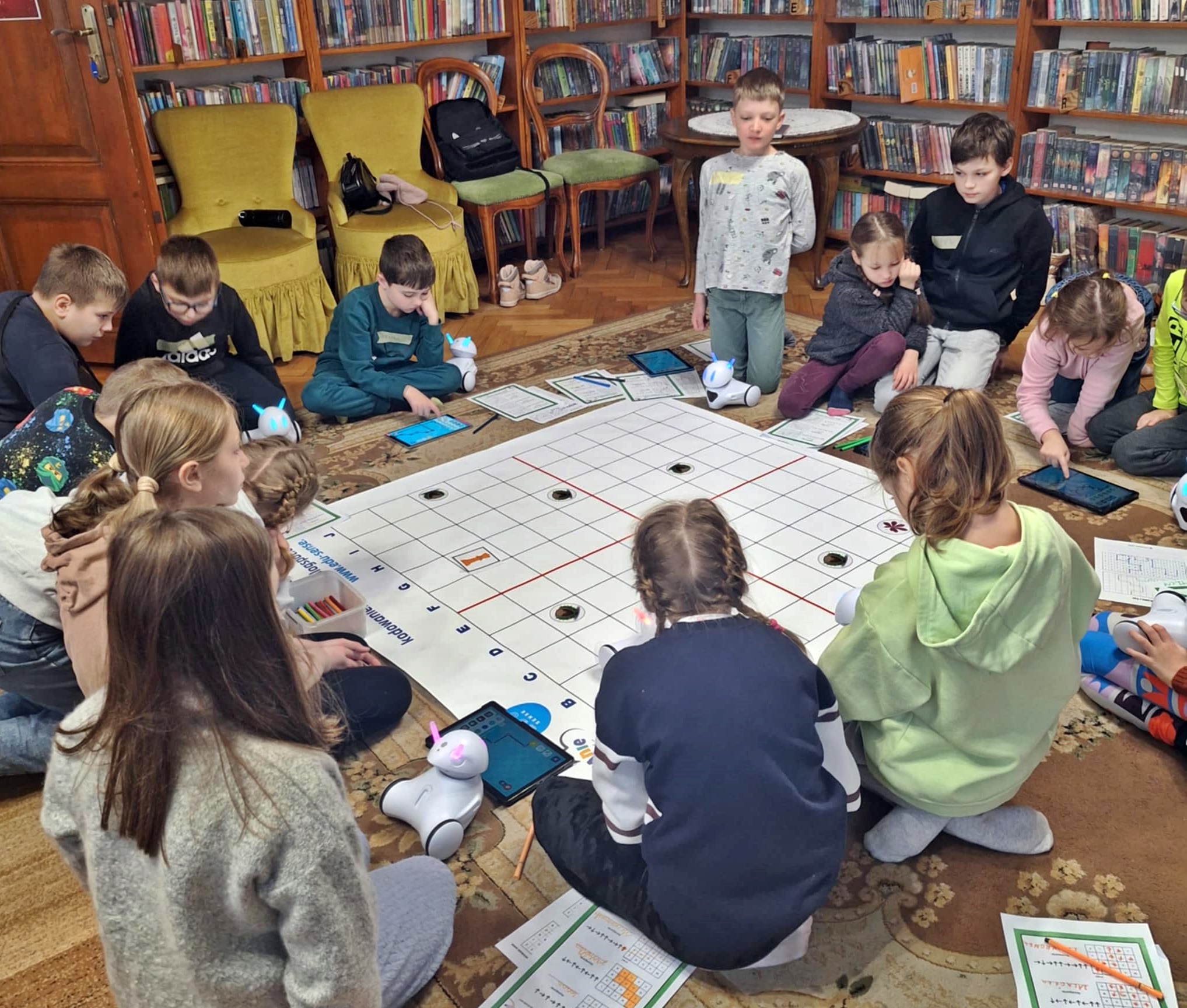 Na dywanie, w kole siedzą dzieci. Przed dziećmi leżą tablety, kartki oraz kilka robotów Photonów. Pośrodku koła leży mata do programowania. Na kilku kwadratach maty programującej położone są obrazki. 
