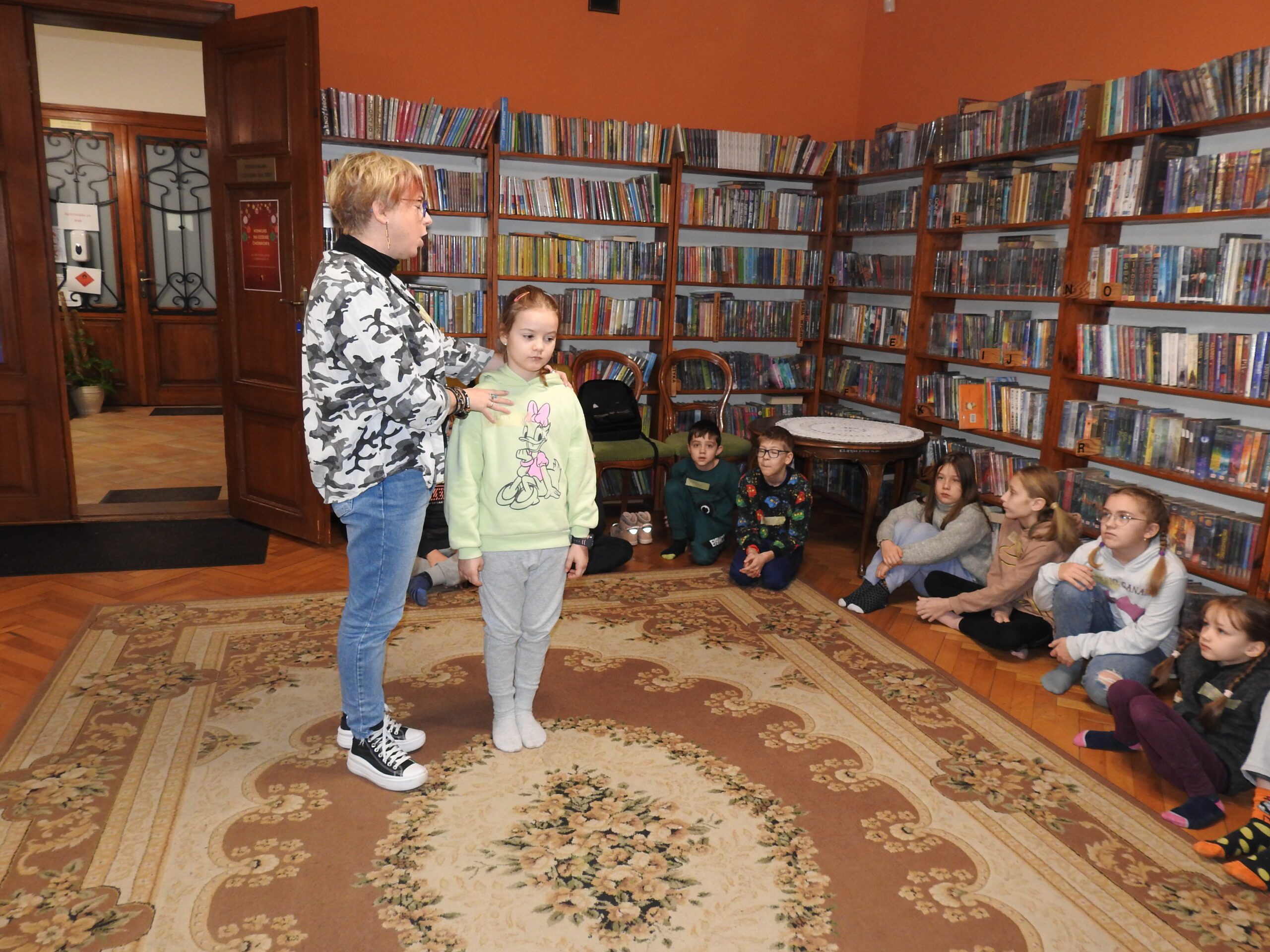 Na dywanie w kole siedzą dzieci. Pośrodku stoi kobieta oraz dziewczynka. Kobieta trzyma dziewczynkę za ramiona. 